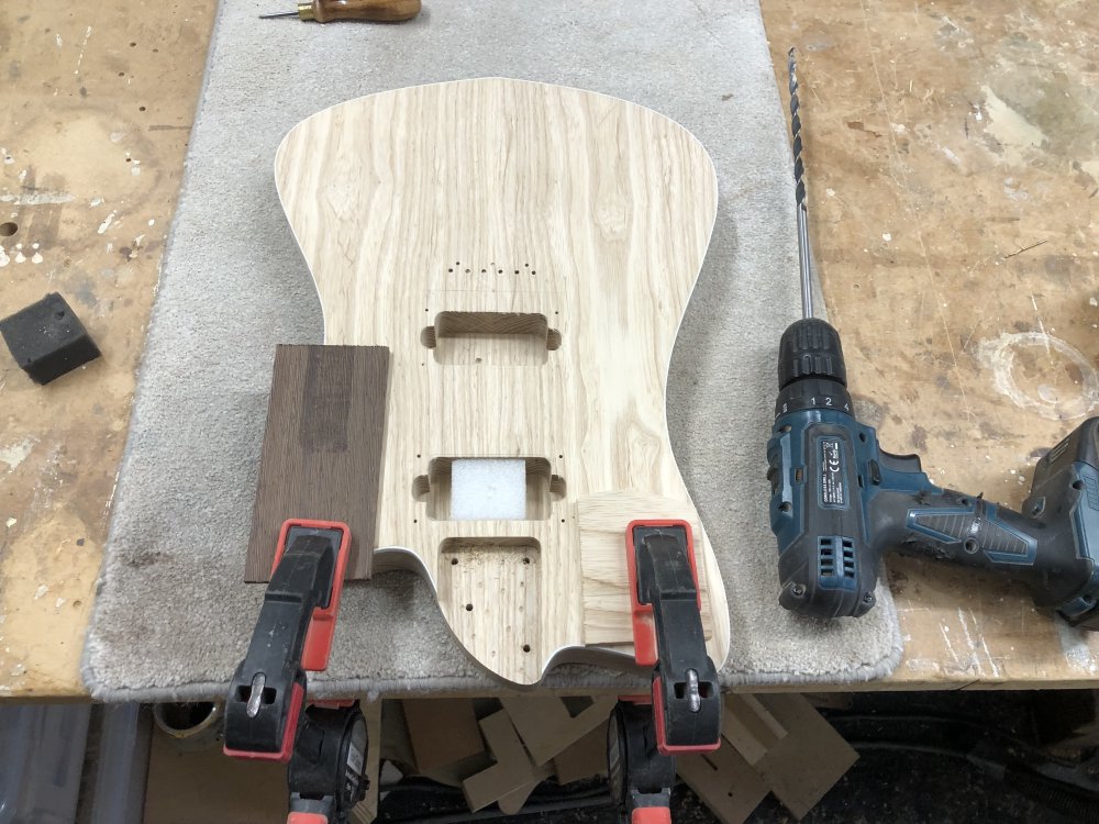 An in progress guitar body sits clamped to the workbench. There are two pickup-cavities, and the one closes to the neck is stuffed with a polystyrene block. Beside the body is a drill in which is an 8mm bit that is about 20cm long.