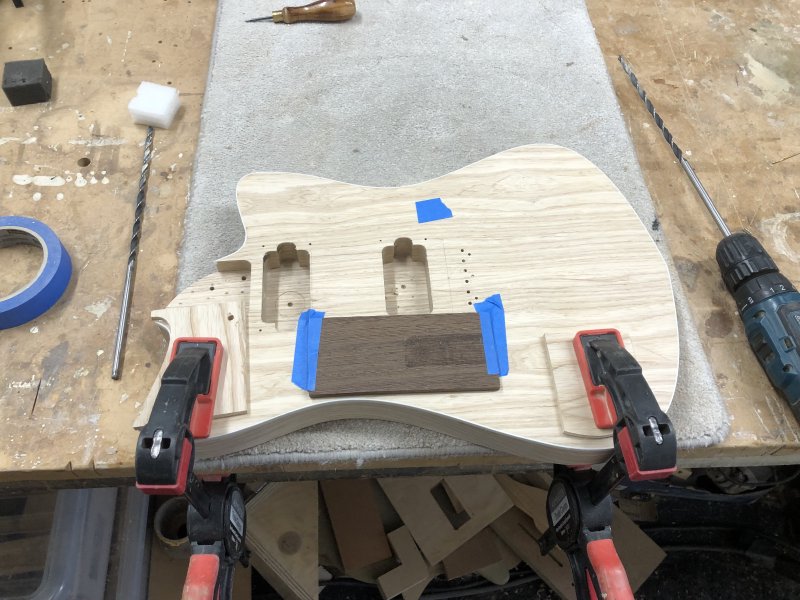 The same guitar body as before, but now it's clamped long edge on, and on the top near the bridge pickup cavity is taped a thin block of wood on the near side, and there's a bit of blue masking tape on the far side.