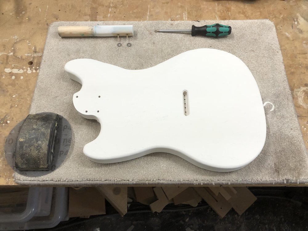 A guitar body covered in white primer sits face down on carpet matt on the workbench.