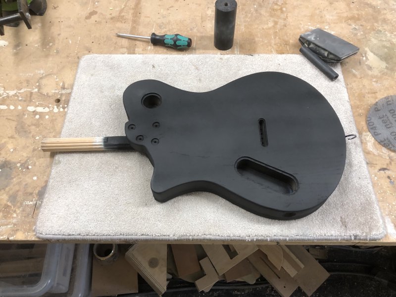 A guitar body covered in black primer sits face down on carpet matt on the workbench.