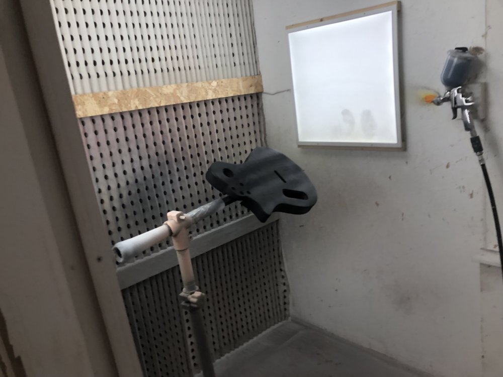 The body of Verkstaden sits mounted in clamp at chest height in the spray-booth, next to a light panel, and on the wall is hung the spray-gun with black primer in it connected to the air-hose.