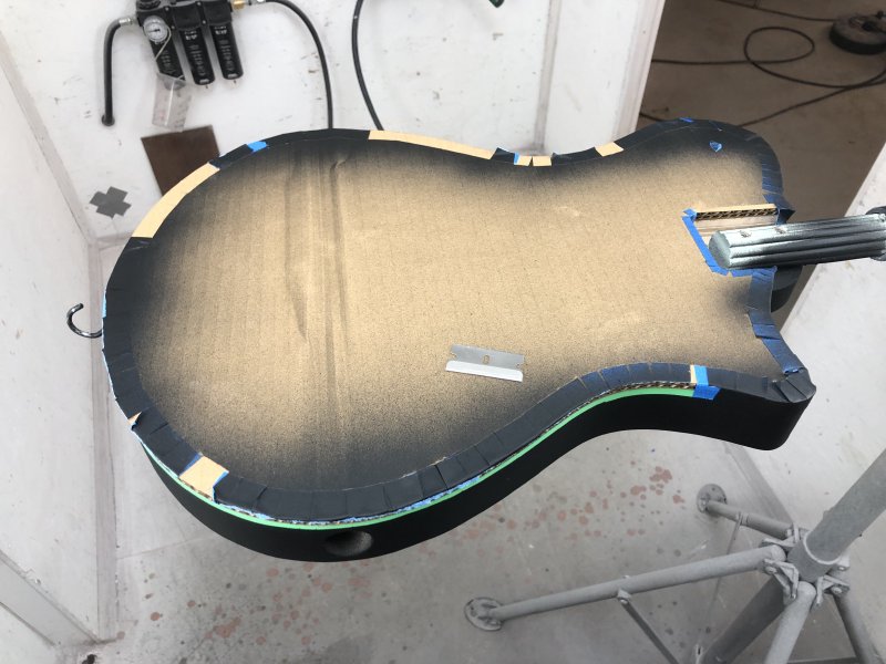 A guitar body is in a clamp, with the front face up. The face has taped to it some cardboard, the edges of which are covered with spray paint. On top of the cover sits a razor blade for removing the tape that holds the cardboard to the guitar body.