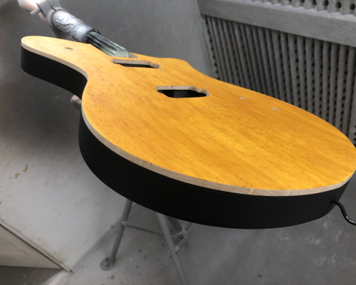 A shot of a guitar body still in a clamp in a spray-booth, showing a yellow stained birds-eye maple front, and then about 5mm of natural wood colour as you go down the side, and the rest of the side is black paint.
