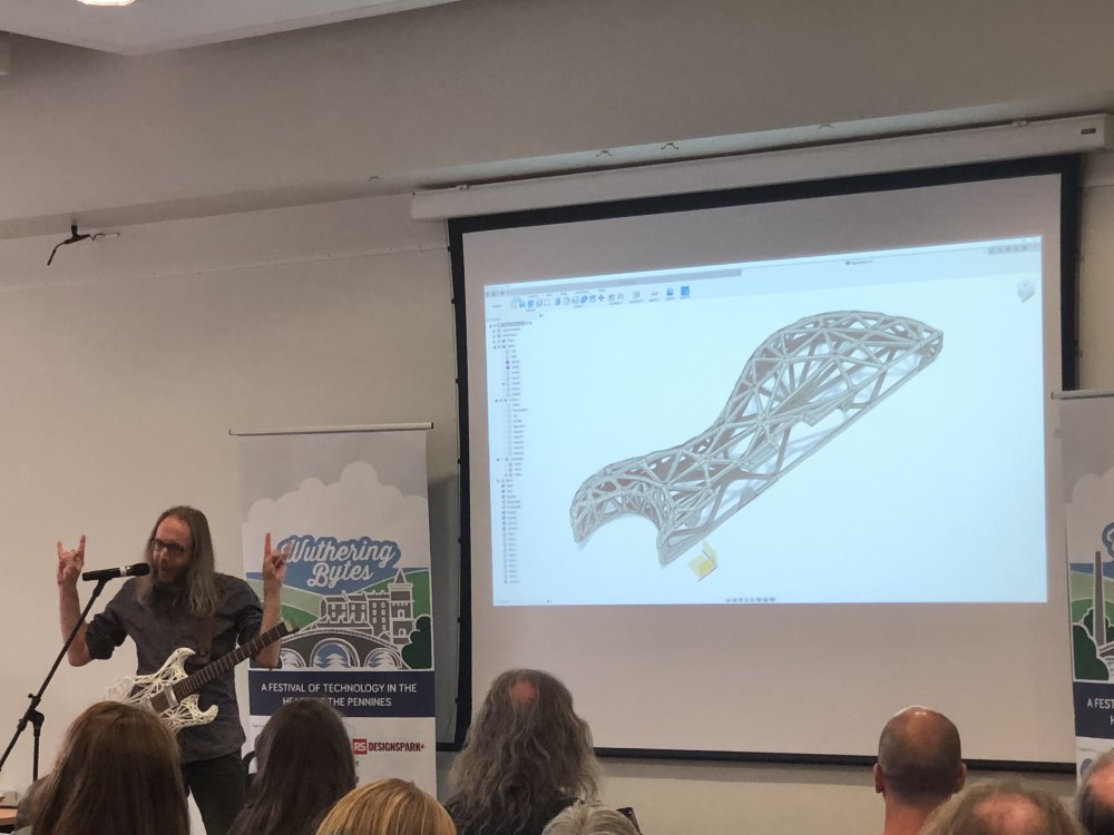 A photo of me talking in front of an audience. I'm stood wearing a guitar on a strap in front of a slide showing a CAD model of the same guitar, and I'm doing the devil's horns gesture with my hands for some reason.