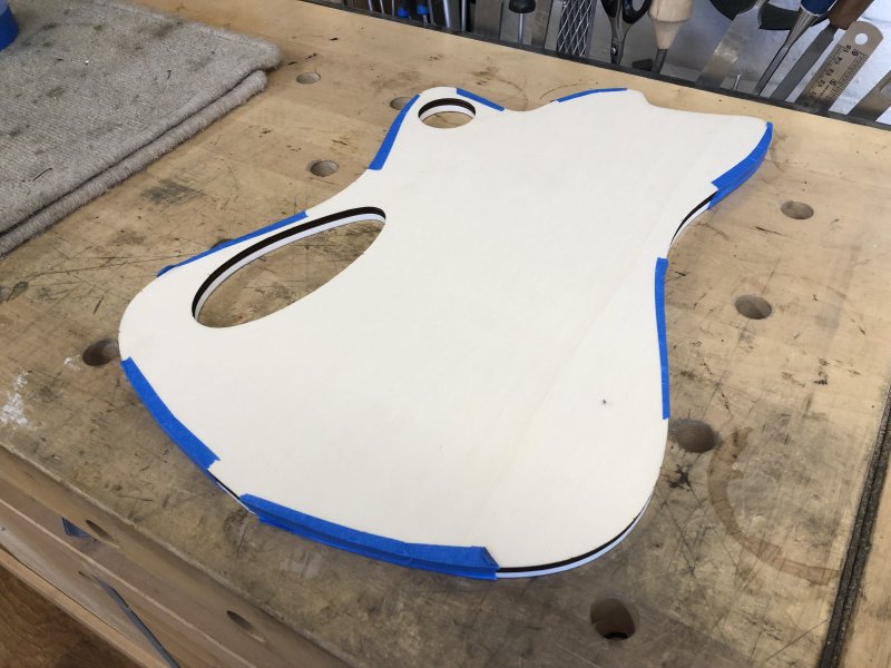 A template sits on the workbench, the shape of a guitar body with two holes in it where the control cavities will be. The template is plywood on top, and white acrylic behind, the two layers held together by blue masking take on all sides.