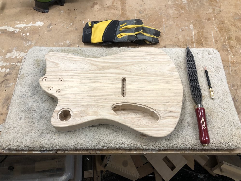 The guitar body sits on a workbench face down, and next to it is a brutal looking saw-toothed rasp, a pencil, and a set of heavy duty gloves. If you were to look closely at the guitar body you could see a pencil sketched outline of where the belly carve will go.