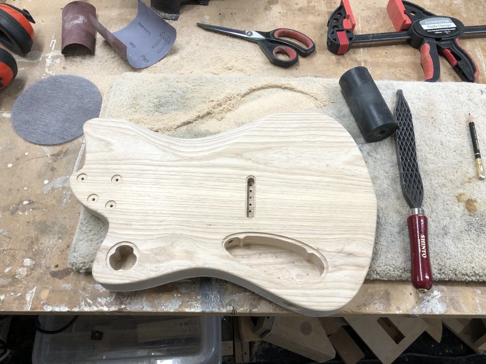 A similar photo to the above of the guitar body face-down on the workbench surrounded by tools, but now with a large amount of wood dust to one side, and a there's now a distinct cut out on the body to allow for someone's belly :)