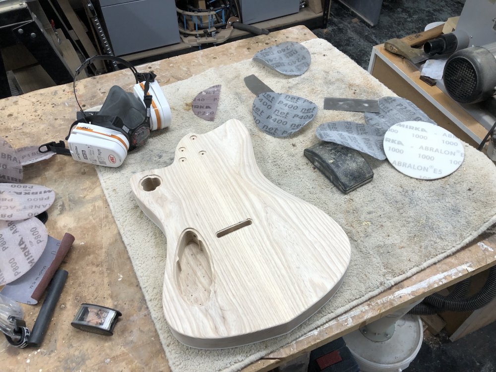 The guitar body sits on the workbench surrounded by sanding disks, wood scrapers, and a respirator mask.