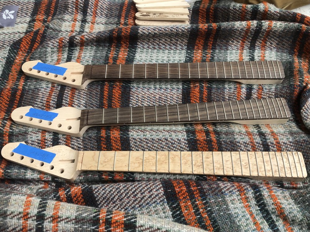 Three guitar necks sit on a tartan rug, two maple with rosewood fretboards and one birds-eye maple with a birds-eye maple fretboard. On the headstock where the logo would normally be is some masking tape with the name of the guitar it belongs to written on it.