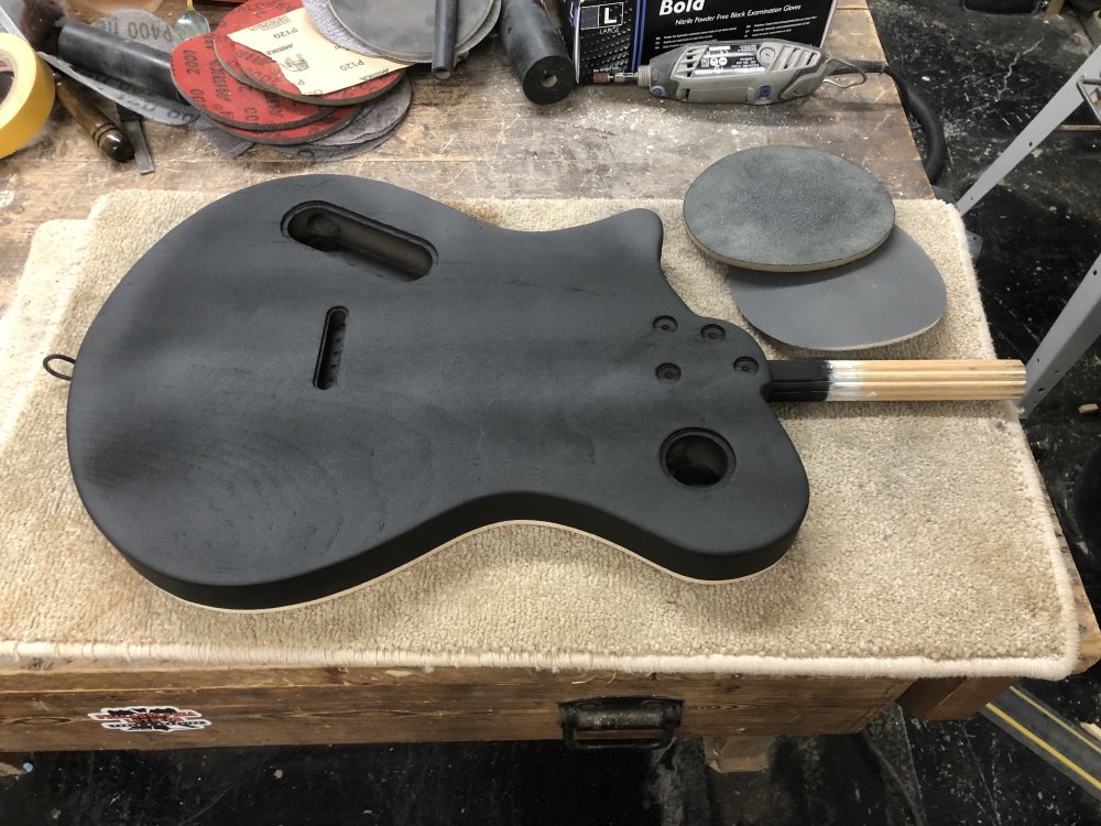 A photo of a guitar body face down on the workshop, covered in black primer, showing streaks from where it has and hasn't been sanded back.
