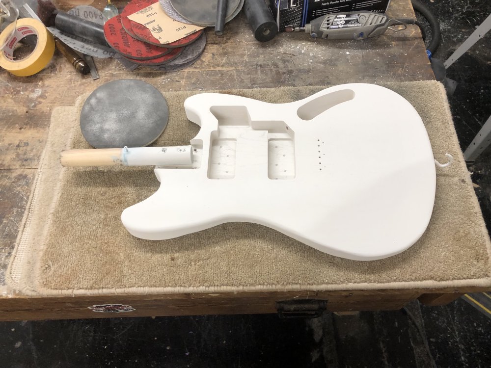 A photo of a Fender Mustang style guitar body on the workbench, coated in white primer, with a sanding pad next to it.