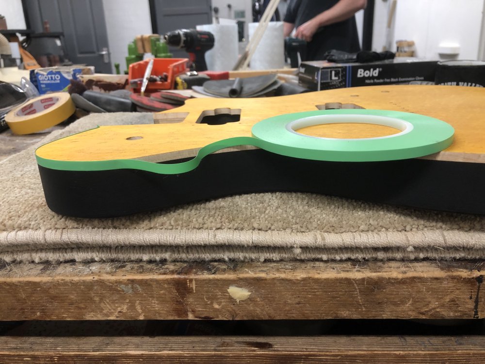 A close-up photo on the side of the guitar body, showing the black primer coated sides, the yellow stained wooden top, and the natural wood fake-binding at the join between the two. A thin piece of green tape is being run around the edge to protect that natural wood line from paint.