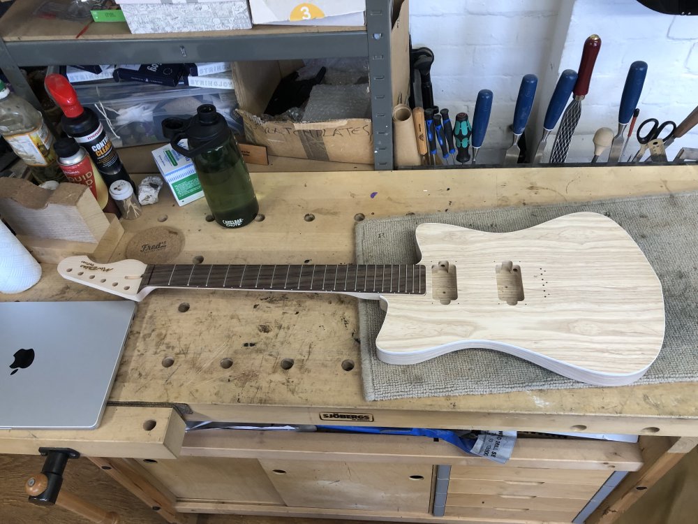 A photo of an unfinished guitar on the workbench. Everything has the right holes etc., but nothing has been coloured or varnished yet. The shape of the guitar is like that of a small Fender offset-guitar with a hint of danelectro styling to it, but slightly pointier.