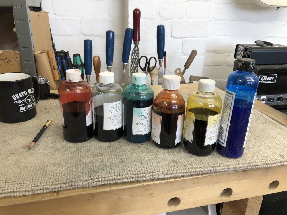 A series of six bottles sit on the workbench, showing red, denim blue, green, orange, yellow, and electric blue wood stains.