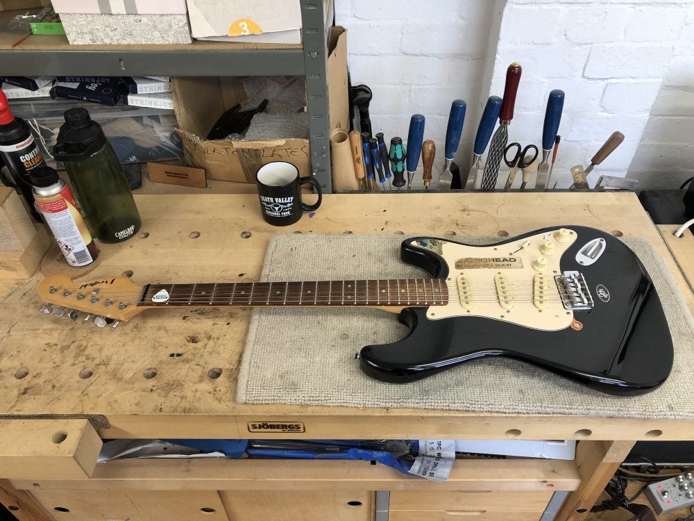 A photo of a black and white Westfield strat-clone guitar sat on the workbench, looking a bit grubby, with faded Radiohead and southpark stickers on it.
