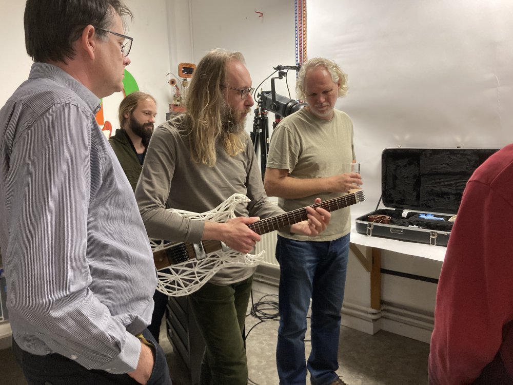 A photo of me holding my Älgen wood and 3D-printed guitar in a cluster of onlookers. I look like I've escaped from a 70s edition of the open university programs.