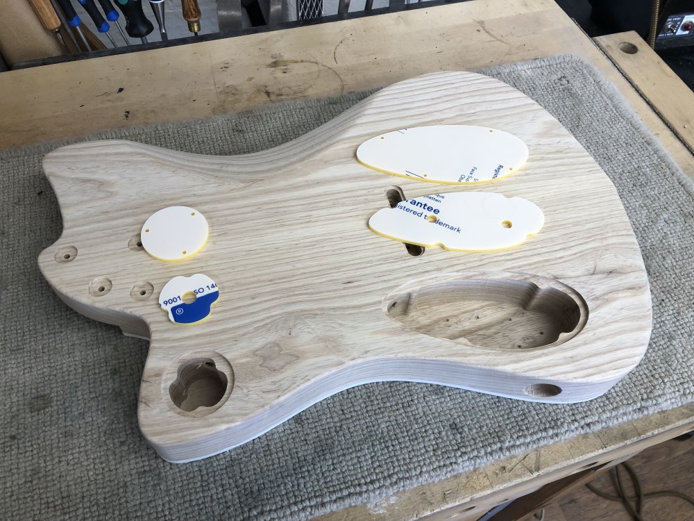 A guitar body sits on the workbench face down, showing a small and large cavity on the back where the electronics parts will be fitted. Next to each cavity are two pieces of laser-cut acrylic: one is a the shape of the bottom of the cavity with holes in it where holes will need to be drilled for the pots, and the other the shape of the lip on the cavity representing the lid.