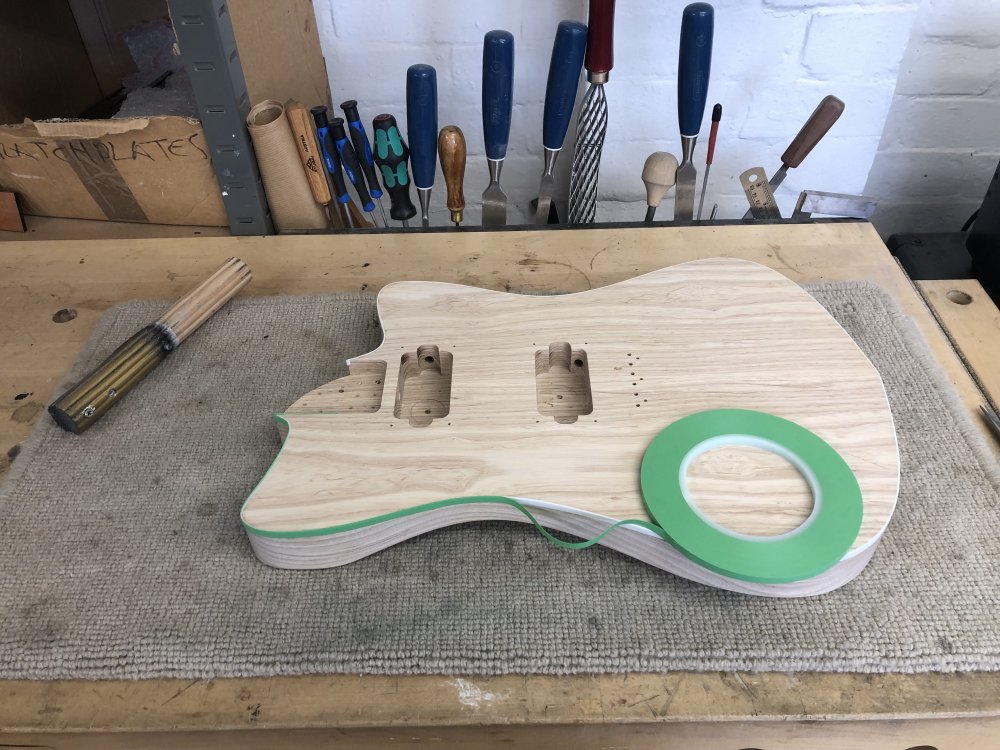 The guitar body its on the workbench, face up this time. Here it is more obvious that there is a white plastic binding that runs around the top edge of the guitar, going about 6mm down the side and 1mm into the top. Over this binding is being placed a thin strip of masking tape.