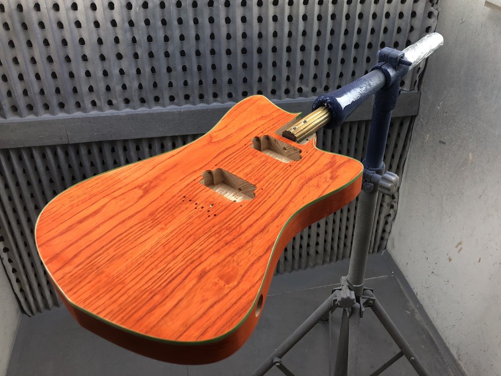 A photo of the guitar, face up in the spray-booth, now having a consistent vivid orange look to the wood, with the grain lines emphasised as they pick up more stain and go darker orange than the rest of the surface.
