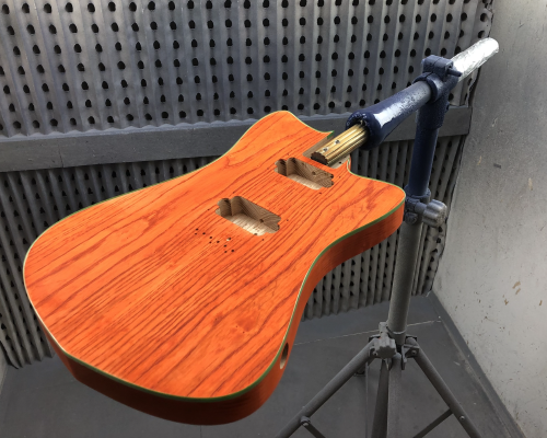 An orange-stained guitar body sits mounted on a jig in a spray booth.