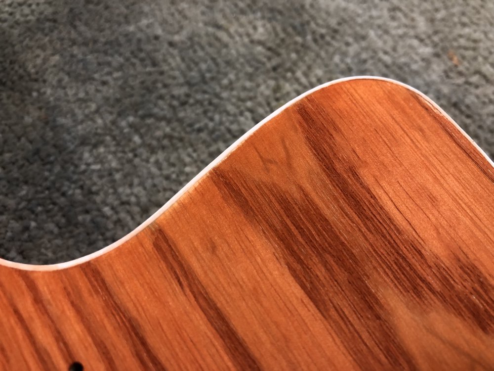 A close-up photo of a corner of the face of an orange stained ash guitar with white plastic binding along the edge. Along an inch or so long section of the edge you can see that the orange stain is notably paler near the binding.