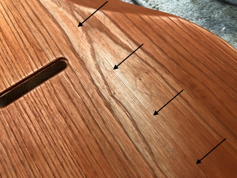 A photo of the back of the guitar body, where a section of the orange stain has been sanded out. A series of arrows drawn on the photo highlight a faint straight line, which might be grain other than it is totally straight, that runs top to bottom along the guitar.