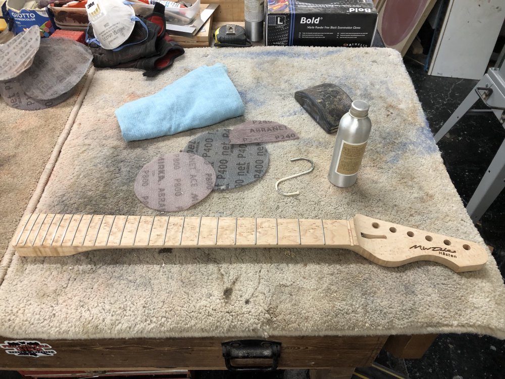 An unfinished birds-eye maple guitar neck sits on a workbench, surrounded by sanding pads, a cloth and a bottle of oil.