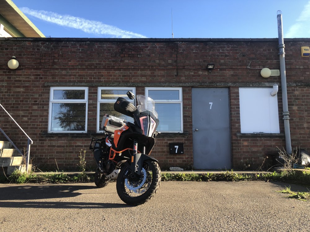 A photo of a large black and orange KTM adventure-motorcycle parked outside a single storey industrial unit, lit up with the sun with blue sky overhead.
