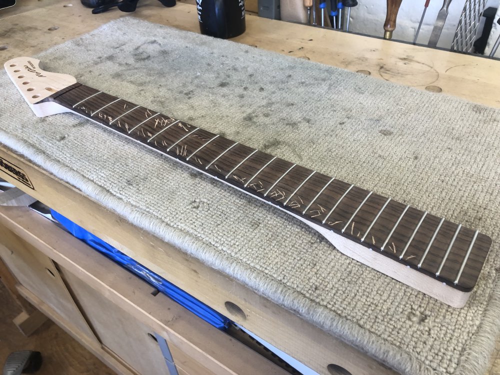 A photo of a maple guitar neck with a rosewood fretboard and frets installed sitting on a workbench. On bits of the fretboard are cross marks in chalk.