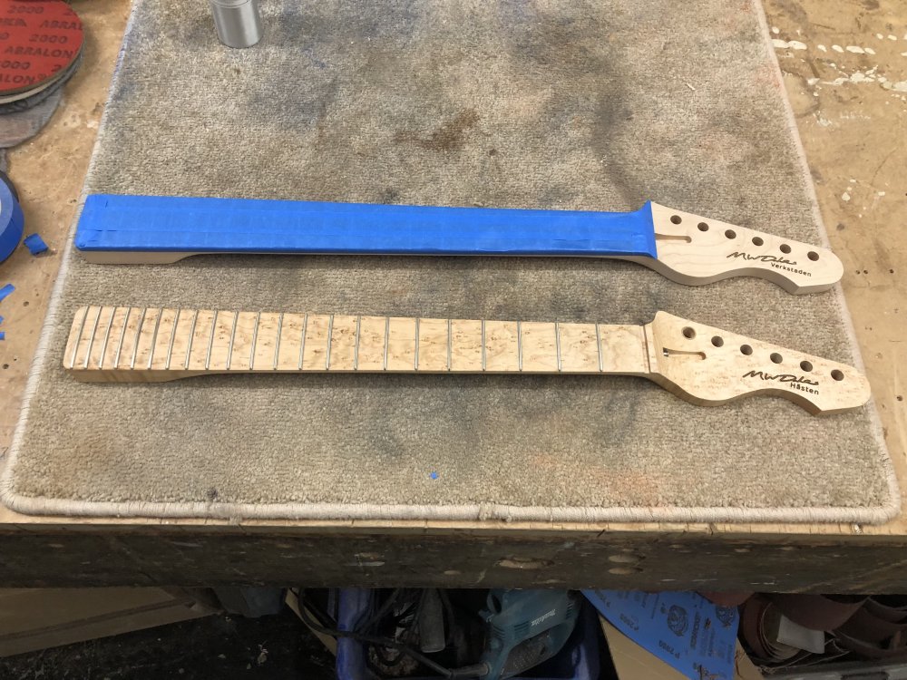 A photo of two guitar necks on the workbench: the one we just saw now has its entire fretboard covered in masking tape, leaving the maple part exposed. Beside it sits the birds-eye maple neck we say being prepped for oiling earlier.