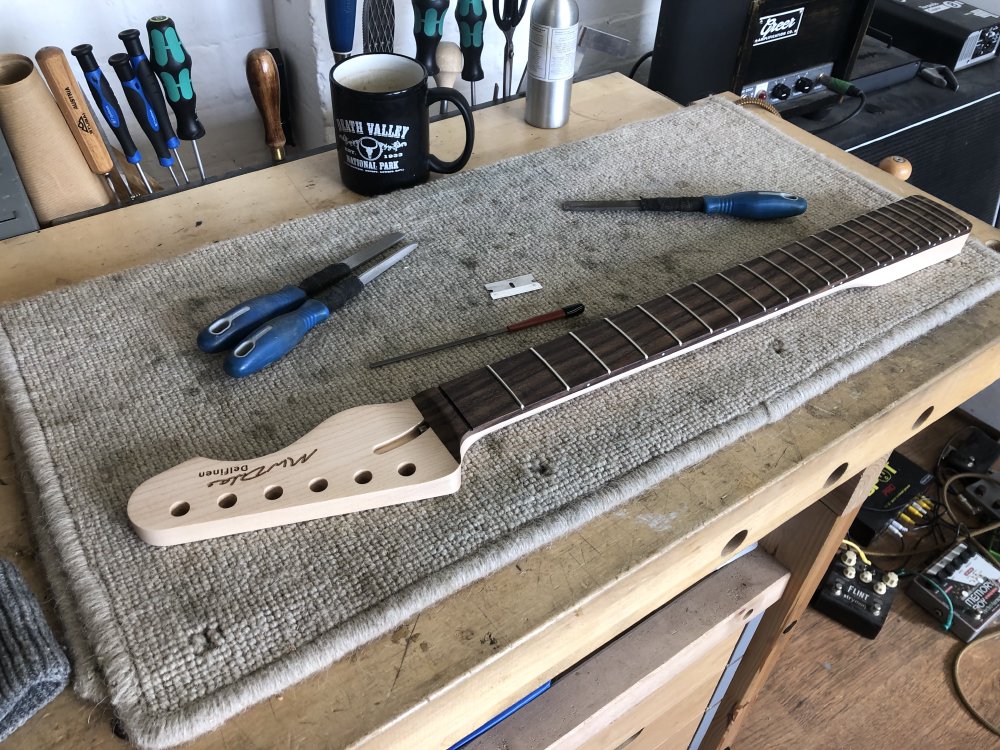 A photo of a guitar neck on a workbench surrounded by files, a razor blade, and a mug. The neck is mostly maple with a rosewood fretboard, and has 22 frets.