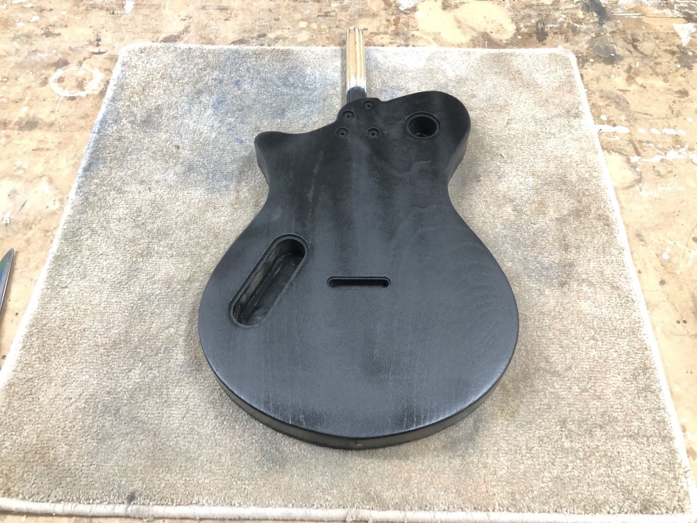 A single-cut style guitar body sits face down on the workbench, with the rear painted black but showing a slight greyness due to being sanded smooth.