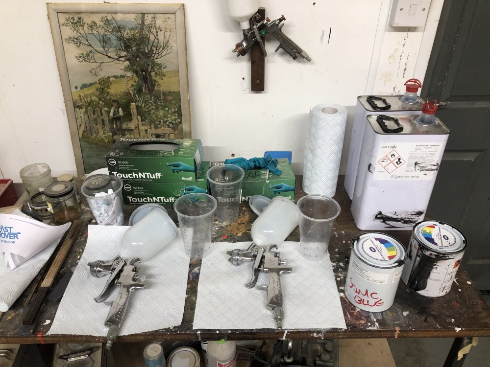 A photo of a paint-spattered table, on which sit two clean spray guns on peices of kitchen roll, next to each is an empty measuing tumbler, and over to the right are two pots of paint, one stainded black and one has 'sonic blue' written on it in sharpie. Behind them are two larger canisters of paint thinners.