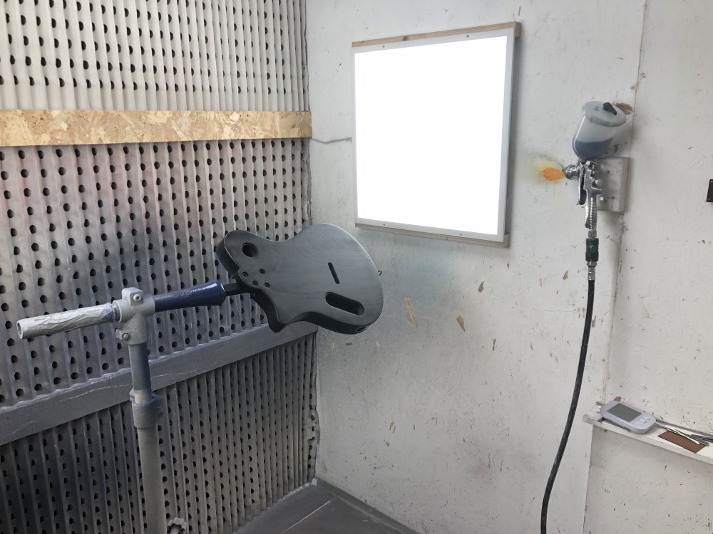 A photo of the single-cut body mounted in the stand in the spray-booth, and on the wall is mounted a spray-gun filled with black paint.