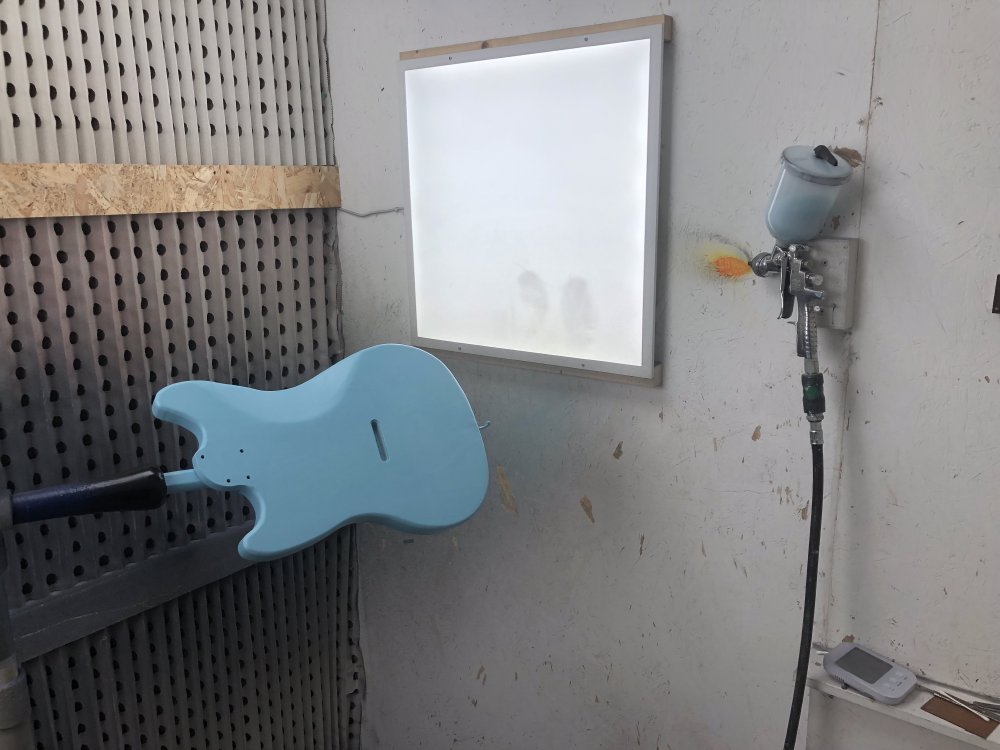 A photo of the blue guitar body mounted on a stand in a spray-booth, and on the wall is mounted a spray-gun with a cup of blue paint loaded.