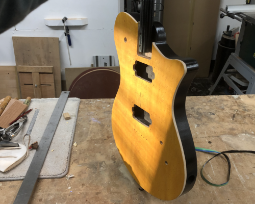 A photo of a guitar body being held over a workbench, caught in the sunlight coming through an unseen window. The face of the body is yellow stained birds-eye maple, and the top 5mm of the side is natural unfinished birds-eye maple, and then the back of the guitar is painted gloss black.