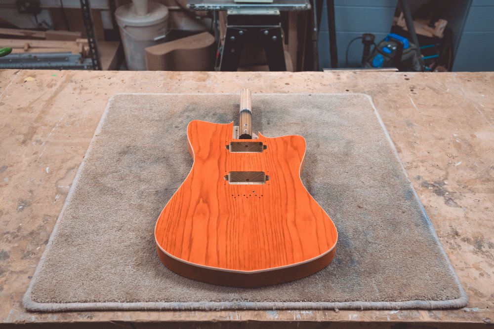 A photo of an offset-style guitar body sat face-up on the workbench, with the wood stained a vibrant orange colour.