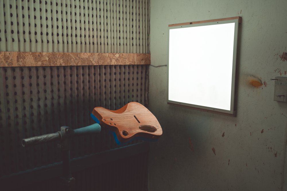 A moodily lit photo of the guitar body on a stand in a spray-booth, lit by a white square light panel. The guitar body was orange and is now sanded to show the wood through the colour.