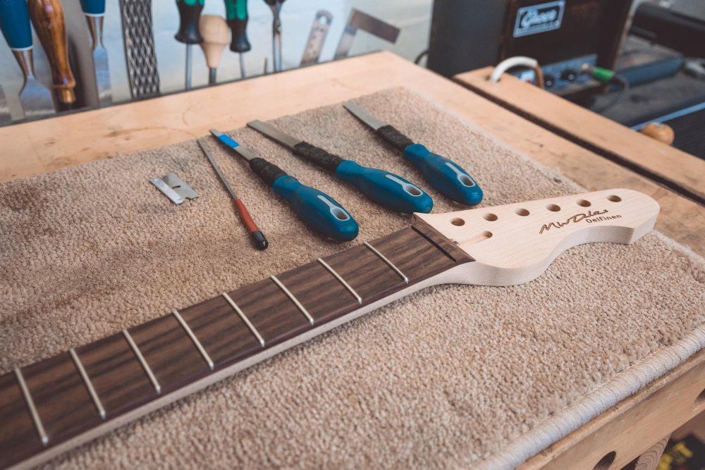 A photo showing the top end of a guitar neck sat on a workbench, next to which sit three large files with different shaped filing ends, a small narrow file, and a razor blade.