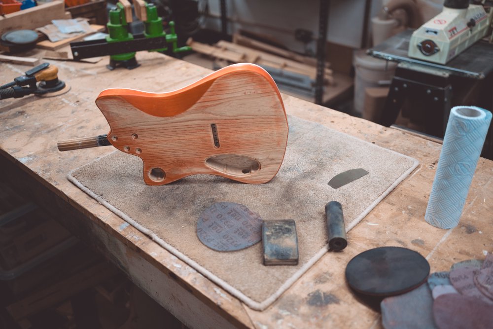 A photo of the guitar body sat on its side on the workbench, surrounded with sanding disks.