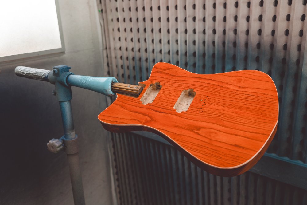 A photo of an orange-stained pointy/offset guitar body on the rack in the spray-booth.