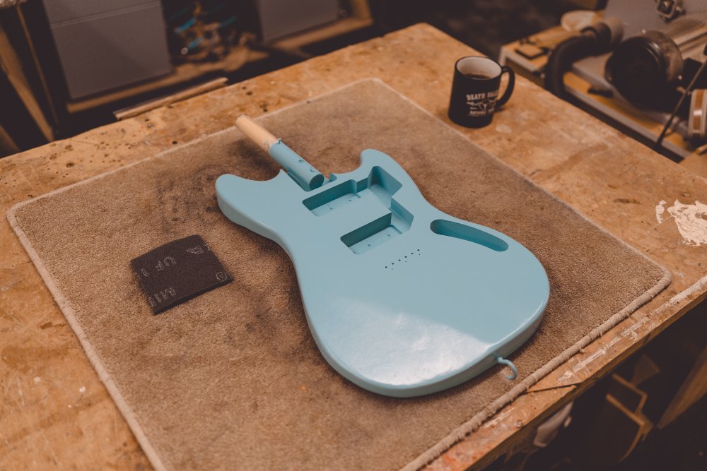 A photo of an offset style guitar body painted a light blue sits on a carpet mat atop a workbench.