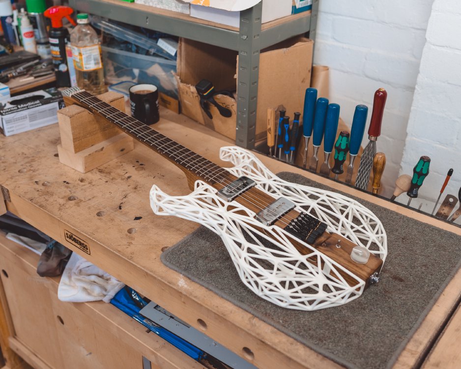 A photo of an unusual looking guitar sat on a workbench: the core of the guitar running from the neck and through the middle of the body area is wood, but the sides of the body are a white 3D-printed lattice work.