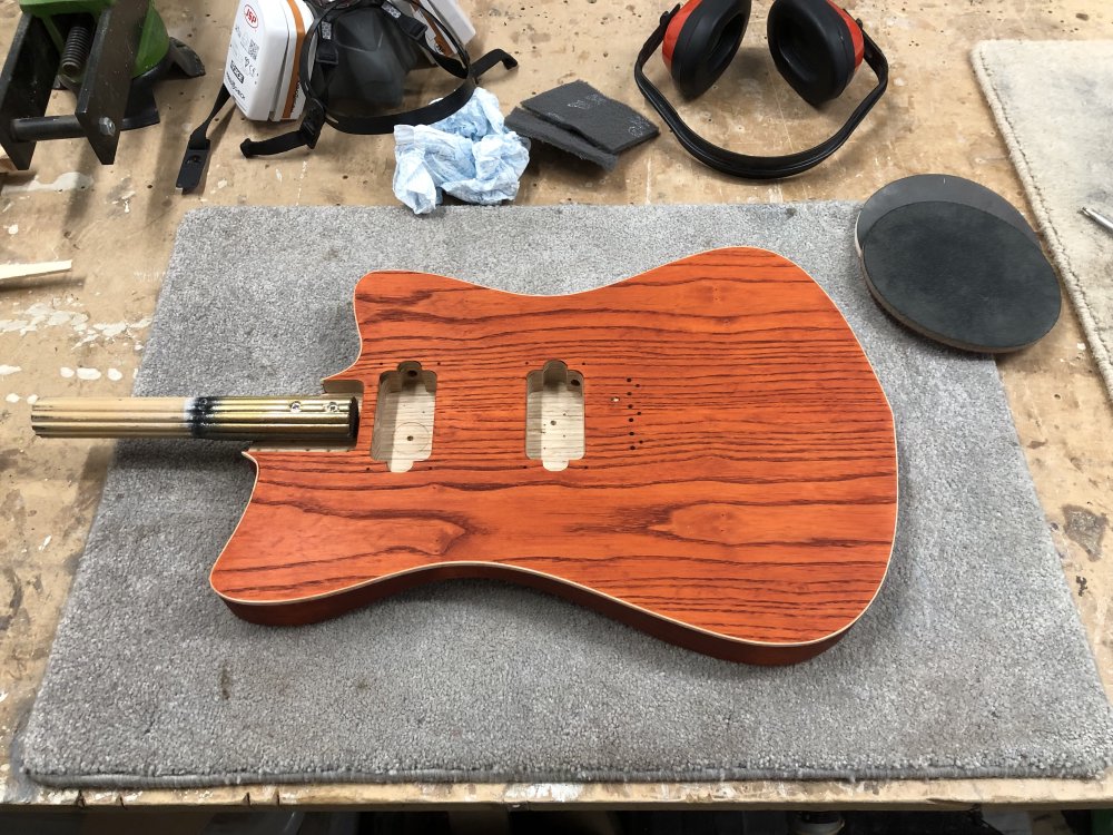 A photo of an offset guitar body on a workbench surrounded by sanding pads and PPE. The body is orange standed swamp-ash with white plastic binding around the top edge, and in the neck pocket is a wood baton screwed in place.
