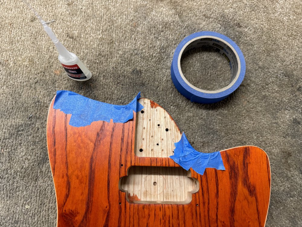 A photo of the guitar body on the workbench, with either side of the neck-pocket there being masking tape strips running from the sides and on top the face of the guitar. Next to the guitar its a small bottle of super-glue and a roll of masking tape.