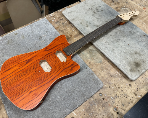 A work in progress guitar sits on the workbench: it has a orange stained swamp-ash body with pockets cut for humbucker sized pickups, it has a maple neck, with a rosewood fretboard and 22 frets. The body shape is a slightly pointy offset style.