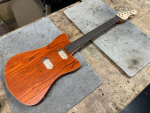 A work in progress guitar sits on the workbench: it has a orange stained swamp-ash body with pockets cut for humbucker sized pickups, it has a maple neck, with a rosewood fretboard and 22 frets. The body shape is a slightly pointy offset style.
