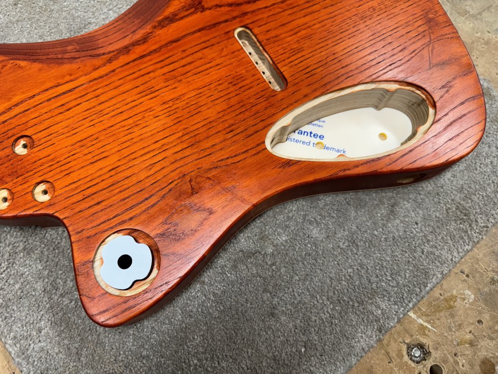 A photo of the orange stained guitar body, only this time it is face down showing the pockets cut into the back where the electronics go. In the larger pocket you can see an acrylic template sits at the bottom showing where to drill two holes, but in the smaller pocket the template sits at the top more like a lid, as it doesn't quite fit the hole.