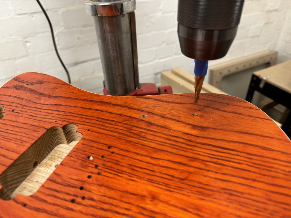 A photo of the guitar body sat on the pillar drill, with the conical drill bit mounted in the chuck, poised above one of the 2mm-ish holes