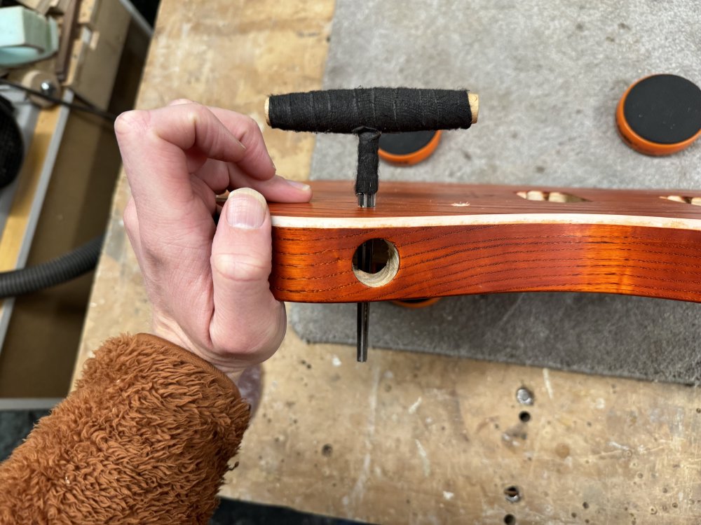 A side on photo of the guitar body, showing a T shaped tool sticking into one of the newly drilled holes. The crossbar on the T is a handle, and the descending part has a sharp edge running down it so it scrapes the wood as you turn the handle.